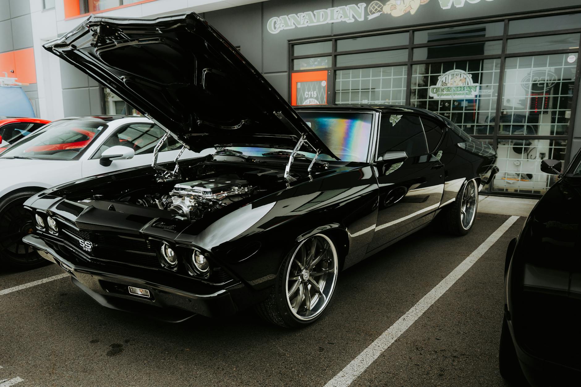 Une voiture de sport noire avec le capot ouvert, mettant en avant un moteur modifié, stationnée sur un parking avec d'autres voitures en arrière-plan.
